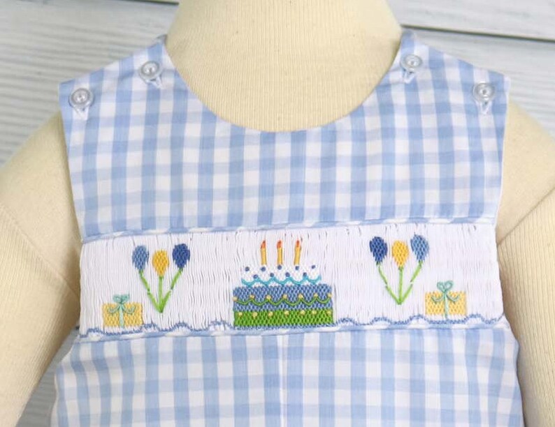May include: A blue and white gingham baby onesie with a white embroidered design of a birthday cake with candles, balloons, and a gift box.