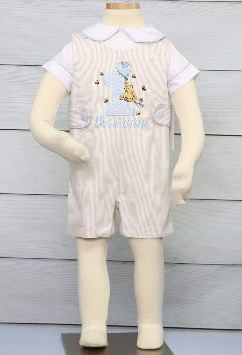 Boys first Birthday Outfit is a light khaki boys romper with side tabs and decorative buttons.  Shown with number 1 and classic winnie the pooh and available with or without monogram.  Matching shirt is also available.