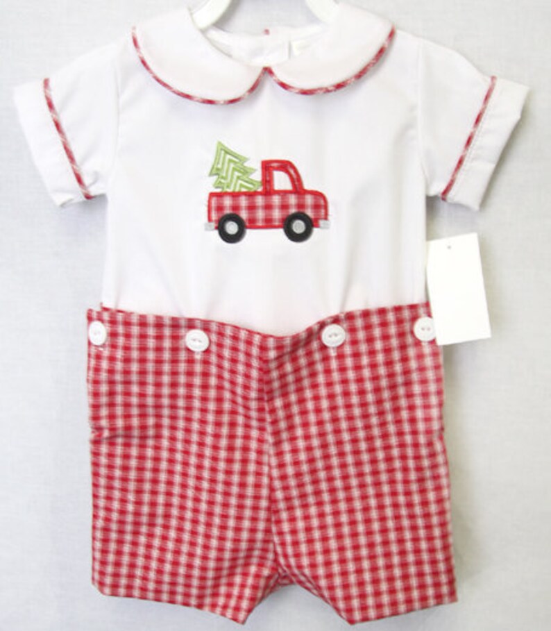 Puede incluir: Un conjunto de beb&eacute; navide&ntilde;o blanco y rojo con un cami&oacute;n rojo y un &aacute;rbol verde. El conjunto presenta un cuello Peter Pan, ribetes rojos y pantalones cortos a cuadros rojos y blancos. El conjunto tiene botones blancos.