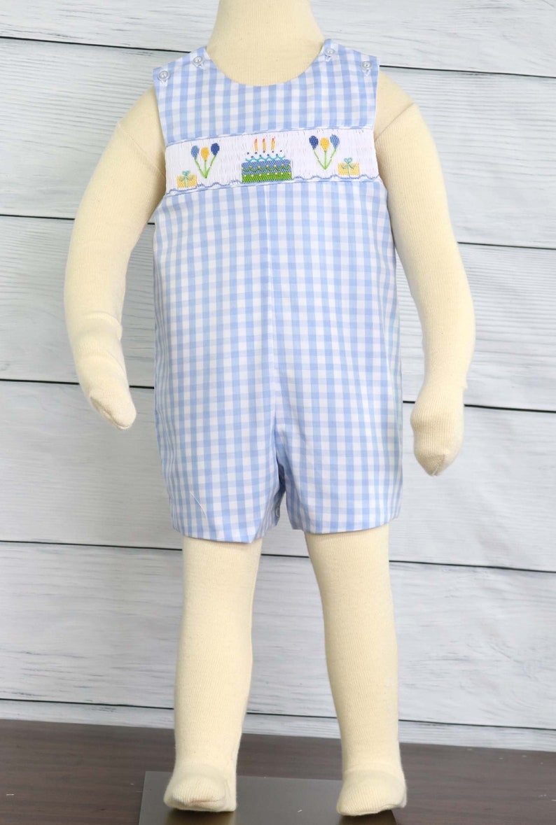 May include: A blue and white gingham baby romper with a birthday cake embroidery design. The cake has candles and balloons.