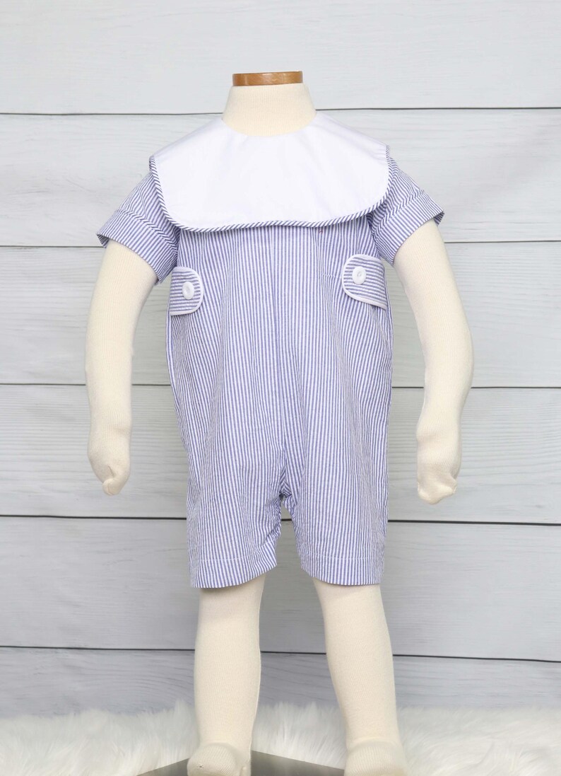May include: Blue and white striped romper with a white collar and short sleeves. The romper has two small pockets with white buttons. The outfit is displayed on a mannequin with beige arms and legs.