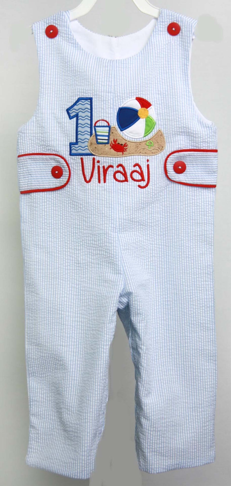 May include: Blue and white striped overalls with a red trim and buttons. The overalls have an embroidered number "1" with a beach scene and the name "Viraaj" on the front.