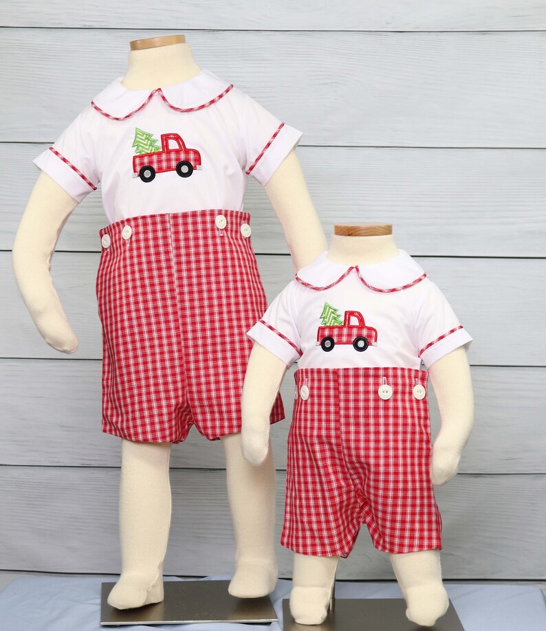 Puede incluir: Dos conjuntos infantiles a juego. Cada conjunto incluye una camisa blanca con cuello con una aplicaci&oacute;n de cami&oacute;n rojo y &aacute;rbol de Navidad, combinada con pantalones cortos a cuadros rojos y blancos. Las camisas tienen ribetes rojos.
