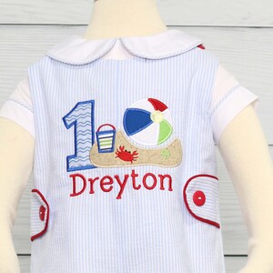 May include: A blue and white striped onesie with a red embroidered number one, a beach ball, a bucket and shovel, and a crab. The onesie also has the name "Dreyton" embroidered on it.