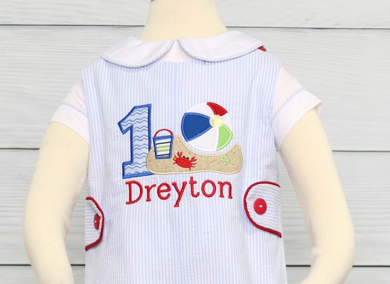 May include: A blue and white striped onesie with a red embroidered number one, a beach ball, a bucket and shovel, and a crab. The onesie also has the name "Dreyton" embroidered on it.