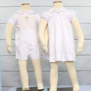 May include: Two white baptism outfits on mannequins. One is a short set with a collared shirt and shorts. The other is a dress. Both have gold cross embroidery and the letters "APN" on the collar.