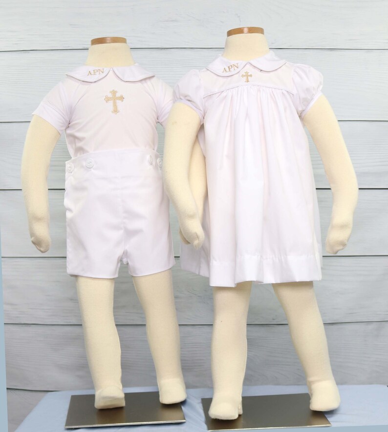 May include: Two white baptism outfits on mannequins. One is a short set with a collared shirt and shorts. The other is a dress. Both have gold cross embroidery and the letters "APN" on the collar.