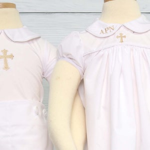 May include: Two white baptism outfits. Each has a gold embroidered cross and the initials "APN". One is a short-sleeved shirt and shorts set, the other a dress with puffed sleeves. The outfits are displayed on mannequins.