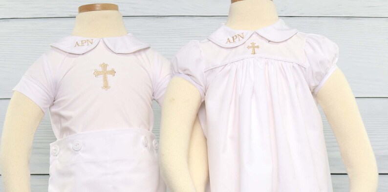 May include: Two white baptism outfits. Each has a gold embroidered cross and the initials "APN". One is a short-sleeved shirt and shorts set, the other a dress with puffed sleeves. The outfits are displayed on mannequins.