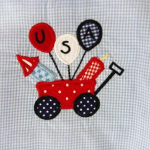 May include: A patriotic applique design featuring a red wagon with white polka dots, carrying fireworks and balloons spelling "USA". The wagon has navy blue wheels with white polka dots. The background is a light blue and white gingham pattern.