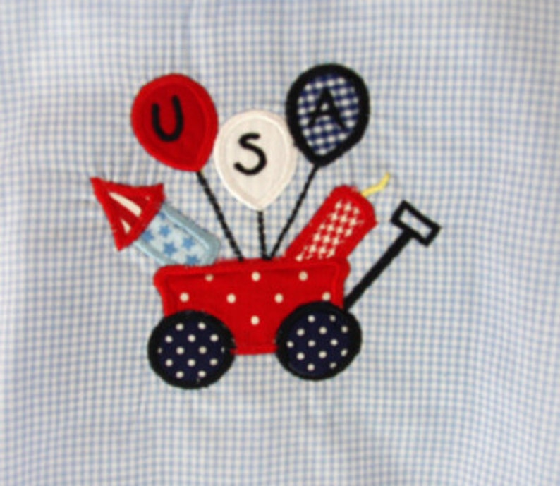 May include: A patriotic applique design featuring a red wagon with white polka dots, carrying fireworks and balloons spelling "USA". The wagon has navy blue wheels with white polka dots. The background is a light blue and white gingham pattern.