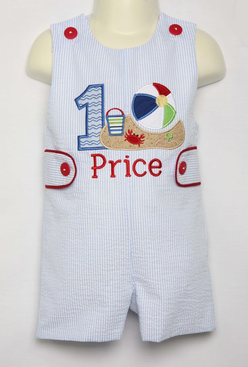 May include: A blue and white striped romper with a red button closure. The romper has an embroidered design of a beach scene with a beach ball, bucket, and crab. The number "1" is also embroidered on the romper. The name "Price" is embroidered below the beach scene.