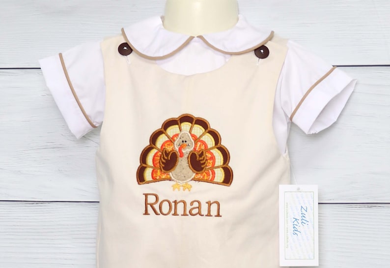 May include: A cream-colored overall shortall featuring an embroidered turkey design and the name Ronan in brown thread. It is worn over a white short-sleeved shirt with a Peter Pan collar and brown piping. A Zuli Kids brand tag is attached.