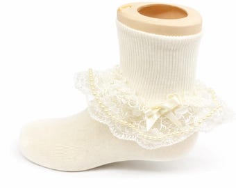 May include: Ivory-colored baby sock with a delicate lace trim, adorned with small pearls and a satin bow. The sock is displayed on a neutral-toned mannequin foot, showcasing its design.