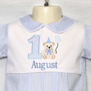 May include: A light blue and white striped romper with a Peter Pan collar. The romper features an embroidered design with the number 1, a teddy bear wearing a party hat, a balloon, and the word "August".