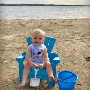 May include: A baby boy wearing a blue and white shirt with a beach theme and the text "Trent" sits in a blue plastic chair on a sandy beach. He is holding a white plastic shovel and a blue bucket is on the sand next to him.