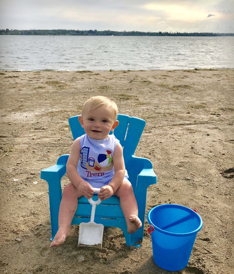 May include: A baby boy wearing a blue and white shirt with a beach theme and the text "Trent" sits in a blue plastic chair on a sandy beach. He is holding a white plastic shovel and a blue bucket is on the sand next to him.