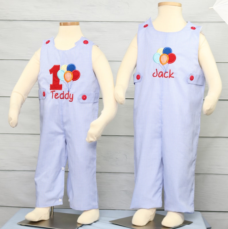May include: Two blue and white gingham overalls with red buttons. One has a red number one and colorful balloons with the name Teddy embroidered on it. The other has colorful balloons and the name Jack embroidered on it.
