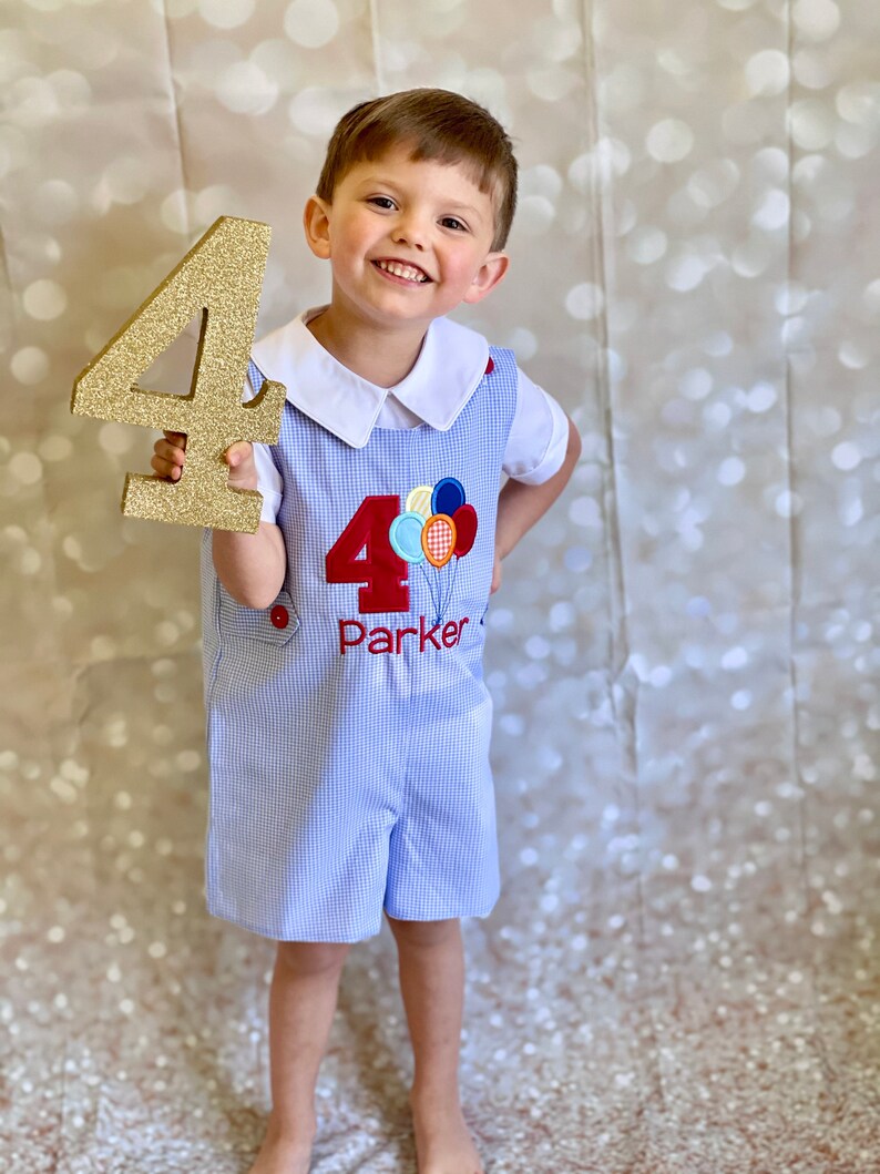 May include: A young boy wearing a blue and white gingham overall outfit with a red embroidered number "4" and the word "Parker" on the front. He is holding a gold glitter number "4" in his right hand and smiling at the camera.