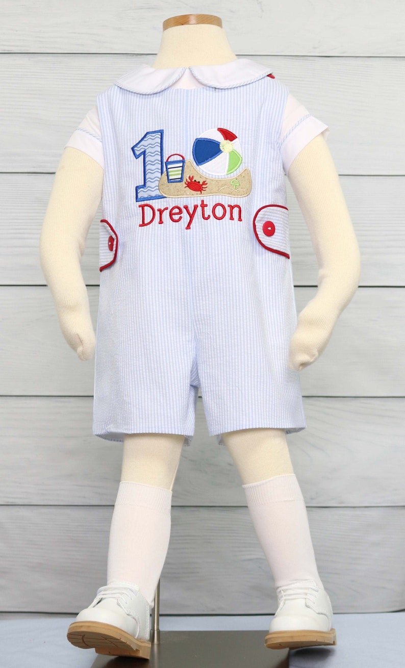 May include: A blue and white striped romper with a white collar and short sleeves. The romper has a red and white embroidered number one with a beach scene and the name "Dreyton" embroidered below. The romper is on a white mannequin.