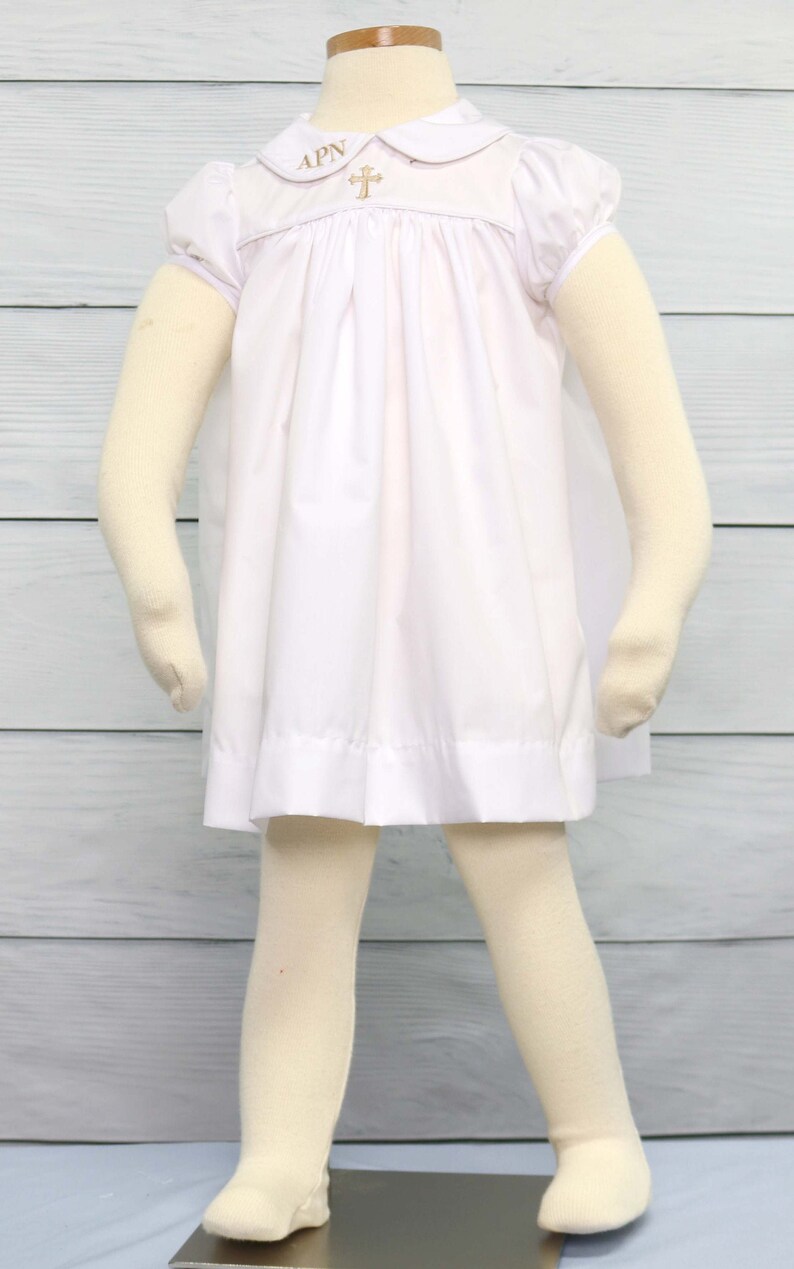 May include: A white baby dress with a Peter Pan collar and short sleeves. The collar has embroidered letters "APN" and a gold cross. The dress is worn with cream-coloured tights and is displayed on a mannequin.