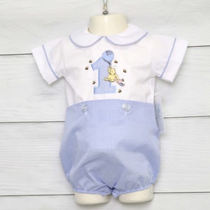 May include: A white short-sleeved onesie with a blue and white gingham pattern on the bottom. The onesie has a white collar with a blue trim and features an embroidered image of Winnie the Pooh holding a blue balloon with the number one and bees around him.