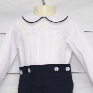 May include: A white shirt with a navy blue trim collar and navy shorts. The shirt has a pleated front and the shorts have white buttons. This outfit is suitable for a special occasion. The measurements are in centimetres.