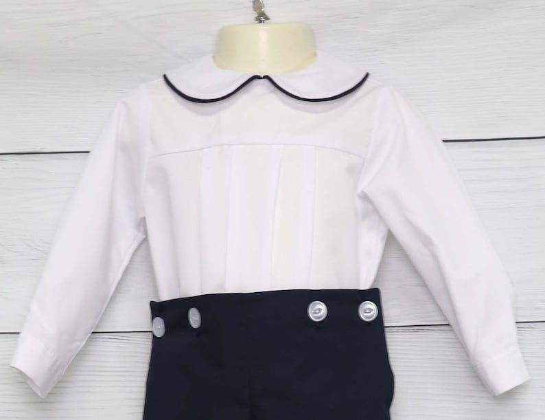 May include: A white shirt with a navy blue trim collar and navy shorts. The shirt has a pleated front and the shorts have white buttons. This outfit is suitable for a special occasion. The measurements are in centimetres.