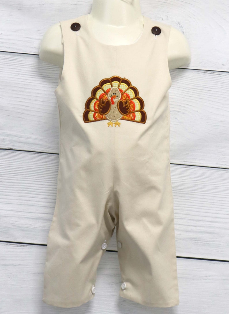May include: A beige sleeveless romper featuring an embroidered turkey design on the chest. The garment has two dark brown buttons at the shoulders and white snap closures along the inner leg seams for easy access. The turkey has brown and orange feathers.