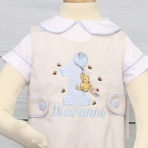 May include: A white and blue striped romper with a Winnie the Pooh embroidery design. The romper has a blue balloon, a number one, and the name "Giovanni" embroidered on it.