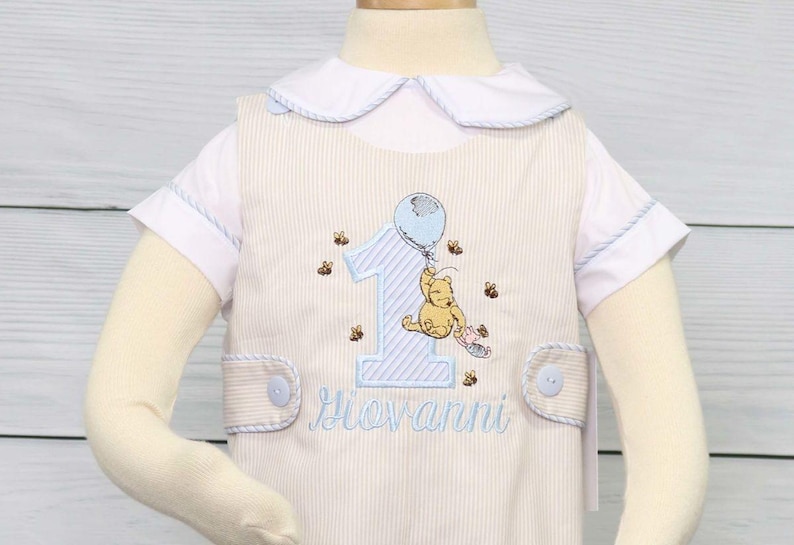 May include: A white and blue striped romper with a Winnie the Pooh embroidery design. The romper has a blue balloon, a number one, and the name "Giovanni" embroidered on it.