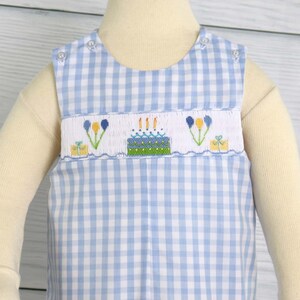 May include: A blue and white gingham sleeveless romper with a birthday cake embroidery design. The embroidery features a cake with candles, presents, and flowers.