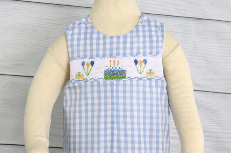 May include: A blue and white gingham sleeveless romper with a birthday cake embroidery design. The embroidery features a cake with candles, presents, and flowers.