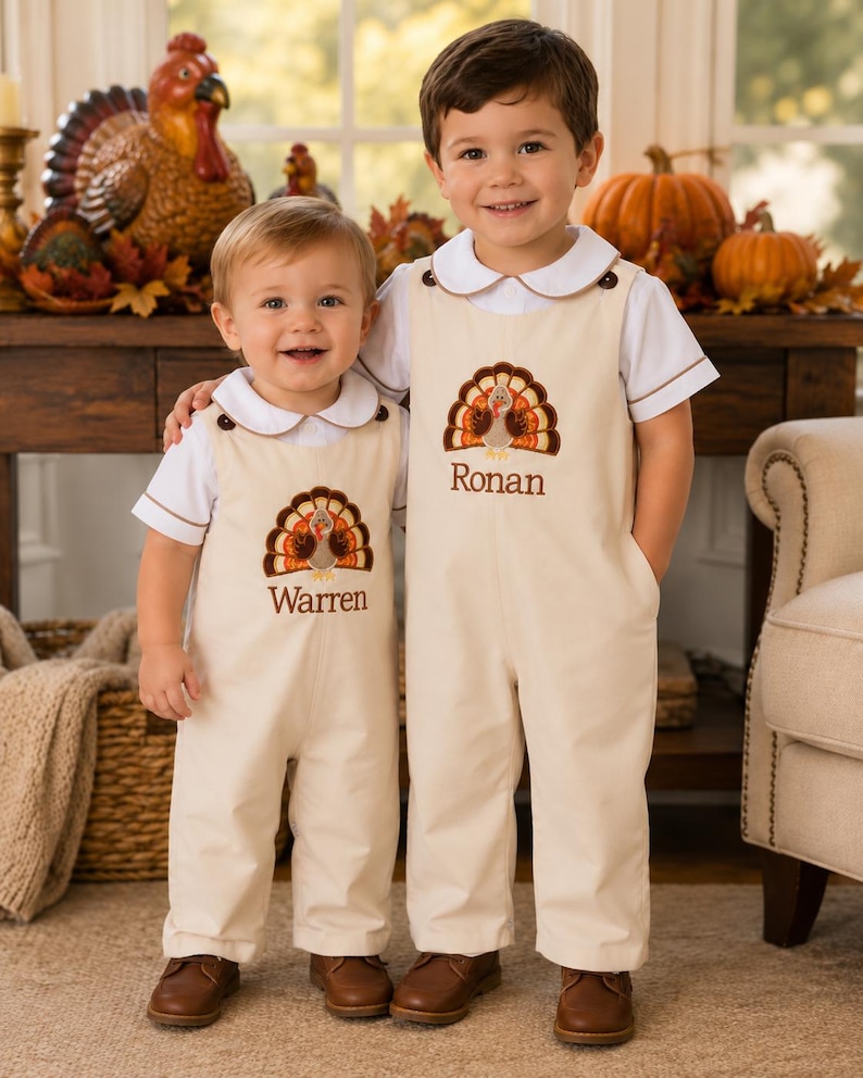 May include: Two beige overalls with white short-sleeved shirts featuring brown piping on the collars and sleeves. Each overall has an embroidered turkey graphic and the names Warren and Ronan stitched in brown thread on the chest area.