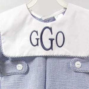 May include: A blue and white striped romper with a white collar. The collar features the embroidered letters "GGO" in navy blue. The romper has short sleeves and two small pockets with white buttons.