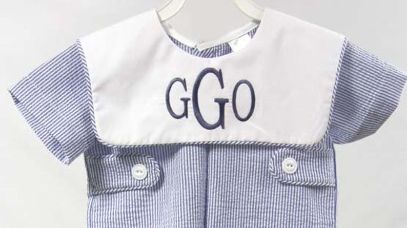 May include: A blue and white striped romper with a white collar. The collar features the embroidered letters "GGO" in navy blue. The romper has short sleeves and two small pockets with white buttons.