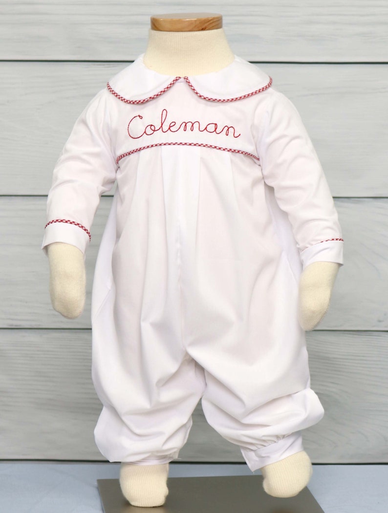 May include: White baby romper with long sleeves and a rounded collar. The collar and cuffs are trimmed with red gingham. The name "Coleman" is embroidered in red script across the chest. The romper has a gathered waist and elasticized ankles.
