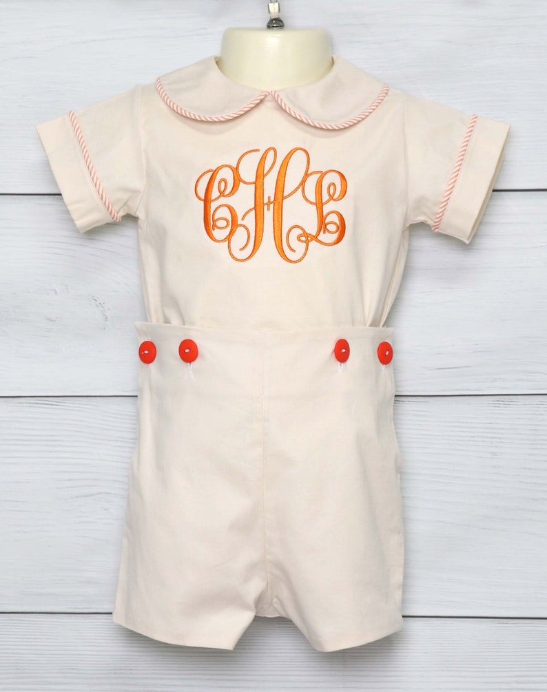May include: Cream-colored romper set with orange monogram and red buttons. The short-sleeved top has a rounded collar and orange trim. The shorts feature three red buttons. This outfit is suitable for a baby or toddler.