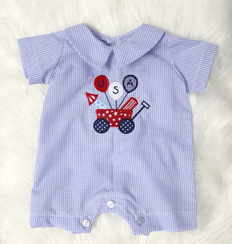 May include: A baby romper in a light blue and white gingham pattern. The romper features a Peter Pan collar and short sleeves. An embroidered design of a red wagon with fireworks and balloons spelling "USA" is on the front.
