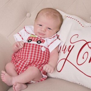 Puede incluir: Un beb&eacute; con un pelele a cuadros rojos y blancos con cuello blanco y un dise&ntilde;o de cami&oacute;n de bomberos. El beb&eacute; est&aacute; acostado sobre una almohada blanca con la inscripci&oacute;n "Merry Christmas" en rojo.
