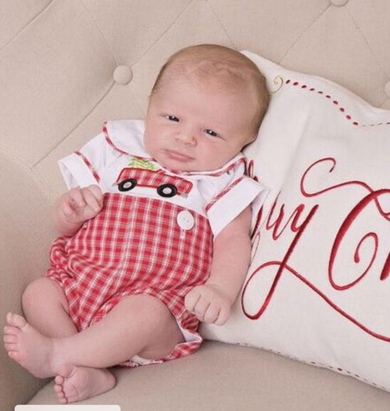 Puede incluir: Un beb&eacute; con un pelele a cuadros rojos y blancos con cuello blanco y un dise&ntilde;o de cami&oacute;n de bomberos. El beb&eacute; est&aacute; acostado sobre una almohada blanca con la inscripci&oacute;n "Merry Christmas" en rojo.