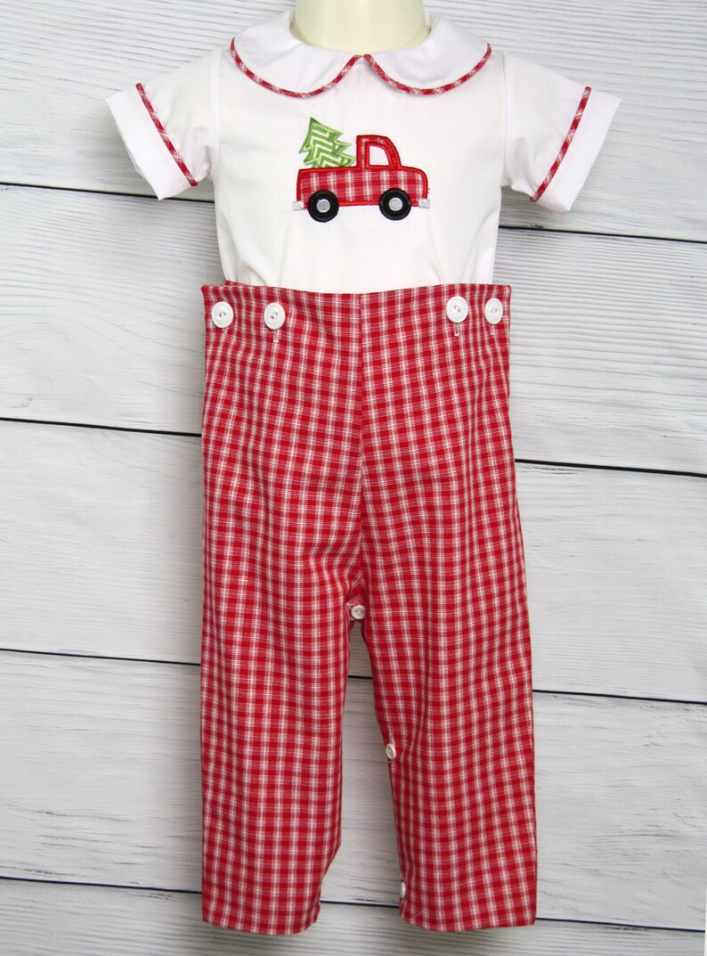 Puede incluir: Un conjunto navide&ntilde;o blanco y rojo para beb&eacute;. La parte superior tiene un cami&oacute;n rojo con un dise&ntilde;o de &aacute;rbol de Navidad. Los pantalones son de cuadros rojos y blancos con botones blancos. La parte superior tiene un cuello blanco con ribete rojo.