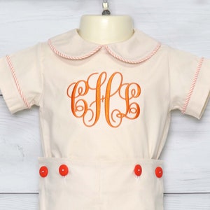 May include: A cream-colored romper set with orange embroidered initials. The outfit features a Peter Pan collar and sleeve and hem trim in a matching orange. The shorts have orange buttons.