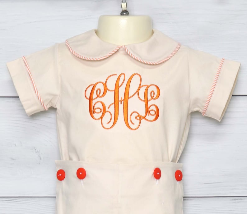 May include: A cream-colored romper set with orange embroidered initials. The outfit features a Peter Pan collar and sleeve and hem trim in a matching orange. The shorts have orange buttons.