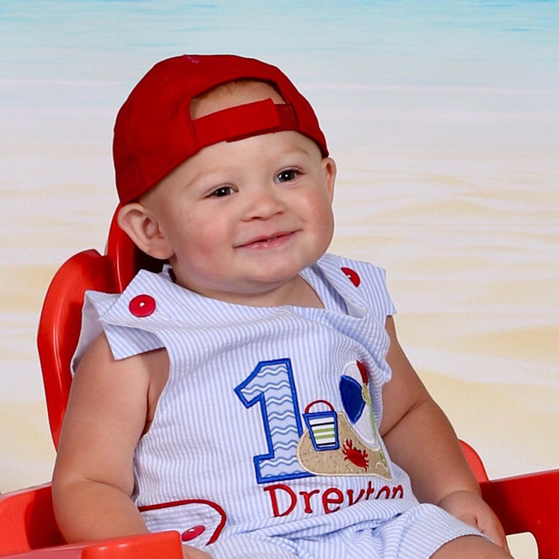 One year birthday outfit is a blue seersucker Jon Jon with side tabs and red buttons and shown with a beach scene and number 1.  Numbers 2, 3, and 4 also availabel.  Can come with or withour baby's first name,