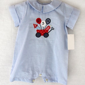 May include: A light blue and white gingham baby romper with a Peter Pan collar and short sleeves. The front features an embroidered red wagon with the letters "USA" and festive balloons and rockets. The romper has button closures.