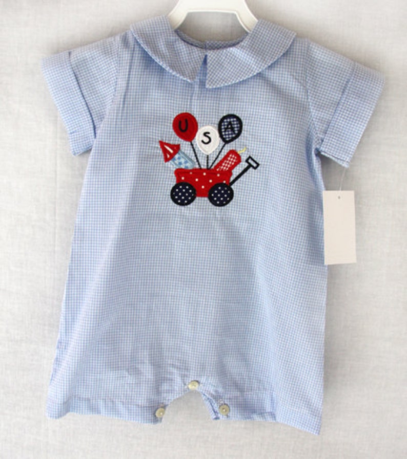 May include: A light blue and white gingham baby romper with a Peter Pan collar and short sleeves. The front features an embroidered red wagon with the letters "USA" and festive balloons and rockets. The romper has button closures.