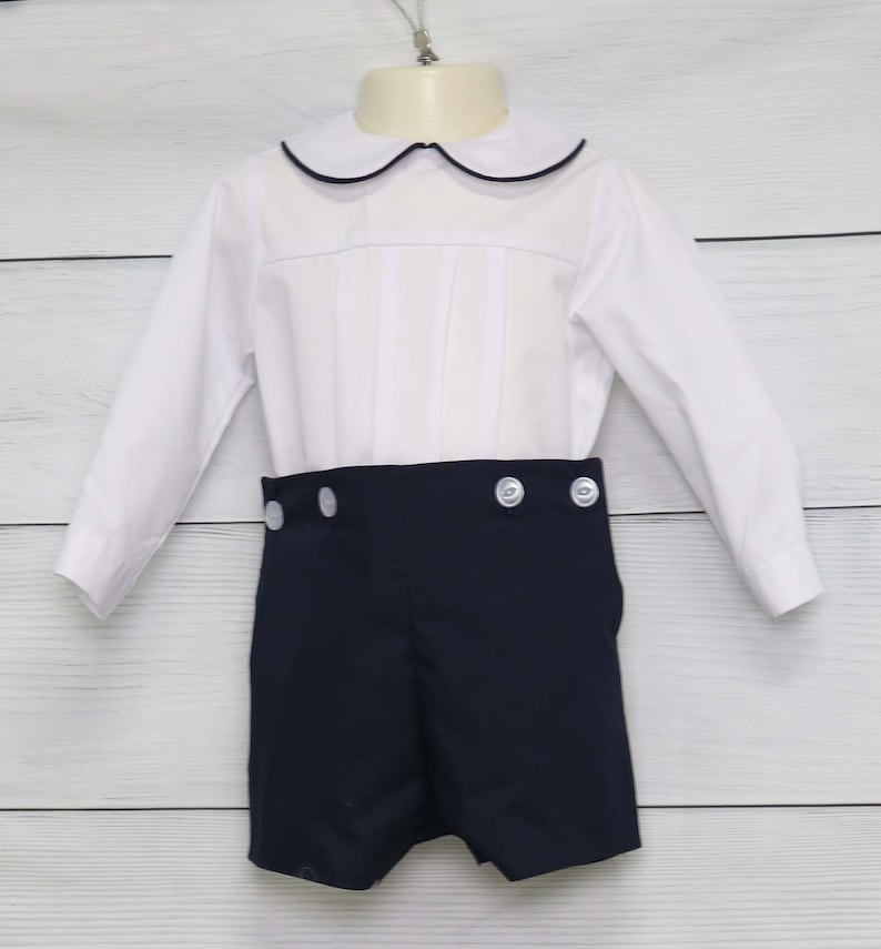 May include: A white shirt with a navy blue collar and pleated front, and navy blue shorts. The shorts have white buttons. This children's outfit is suitable for special occasions.
