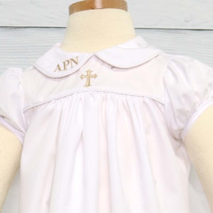 May include: White baby baptism gown with a Peter Pan collar and short, puffed sleeves. The gown features a gold embroidered cross and initials on the bodice. The fabric is soft and gathered, creating a flowing silhouette.