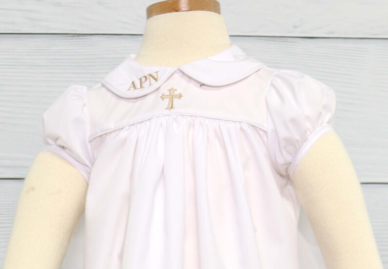 May include: White baby baptism gown with a Peter Pan collar and short, puffed sleeves. The gown features a gold embroidered cross and initials on the bodice. The fabric is soft and gathered, creating a flowing silhouette.